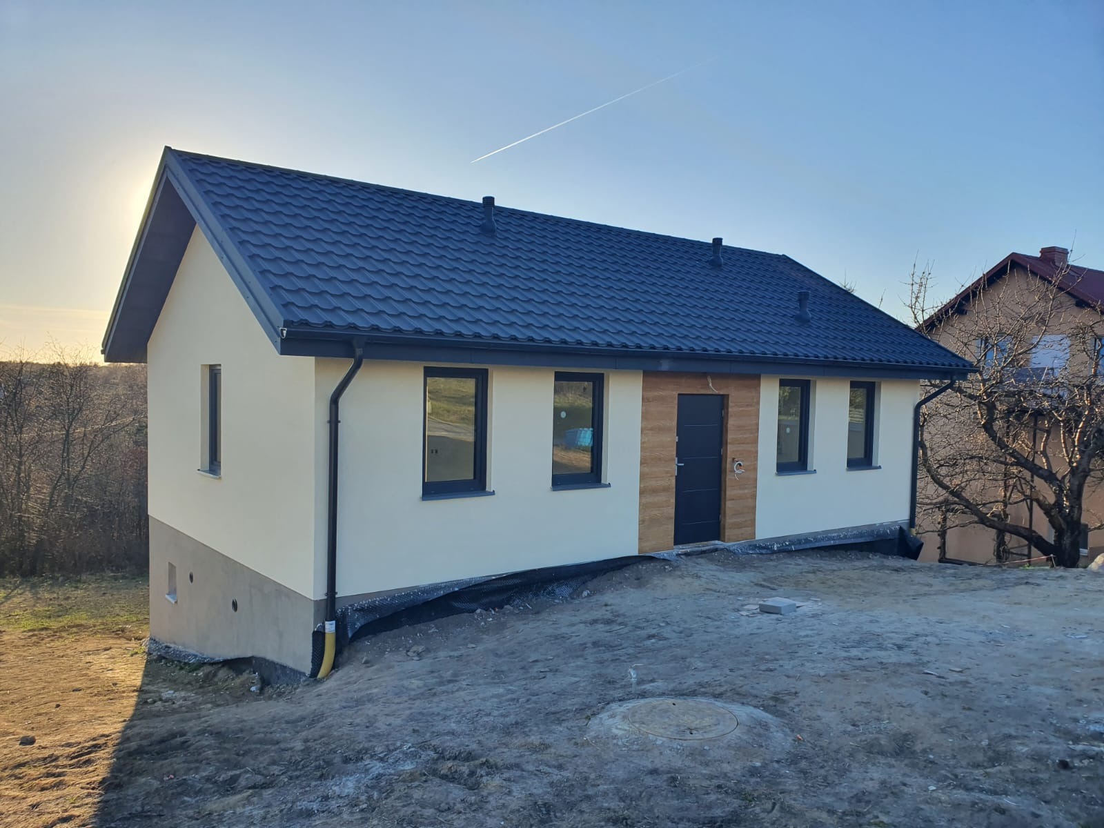 Gotowy dom z ciemną blachodachówką i jasną elewacją, z drewnianymi akcentami przy drzwiach wejściowych. Widoczne czarne okna i rynny. Teren wokół domu jeszcze niezagospodarowany.