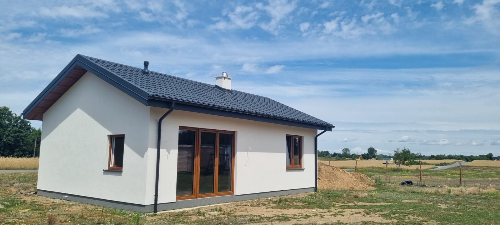 Nowoczesny, parterowy budynek z ciemnym dachem i jasną elewacją, drewniane okna, na działce z widocznym kopcem ziemi. Błękitne niebo z chmurami w tle.