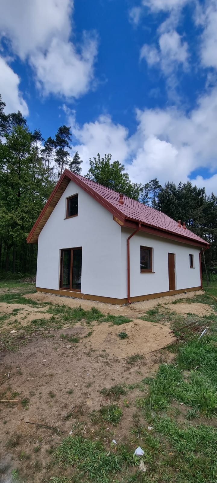Biały dom z czerwonym dachem na tle lasu i błękitnego nieba. Widoczne brązowe okna i drzwi. Dom otoczony ziemią i zielenią.