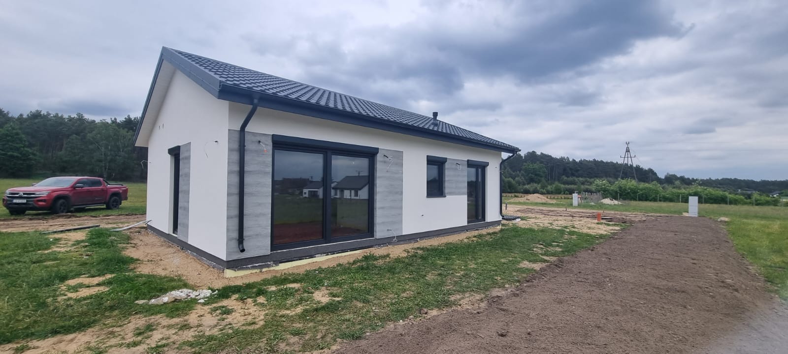 Nowoczesny dom z białymi ścianami i szarymi akcentami, czarną stolarką okienną i ciemnoszarą dachówką. Na posesji stoi czerwony pickup.