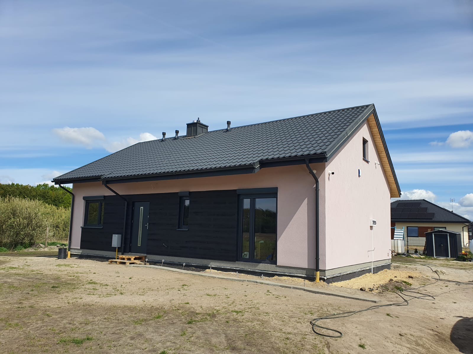 Niskoenergetyczny dom z dachem z blachodachówki, jasnoróżową elewacją i czarną drewnianą fasadą, otoczony piaszczystym terenem i błękitnym niebem.