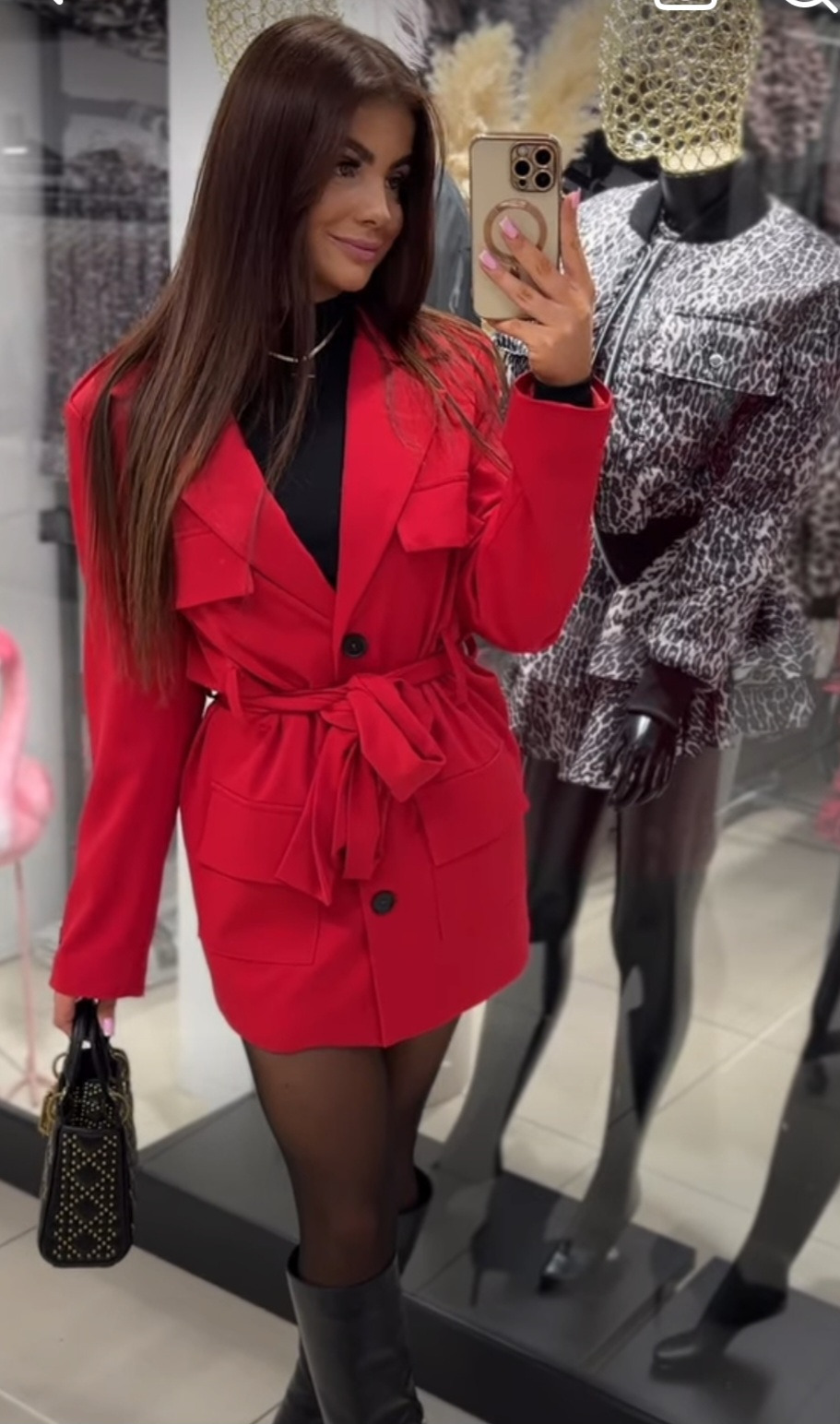 Kobieta robi selfie w czerwonym płaszczu z paskiem, czarnych rajstopach i kozakach, w tle manekin z wzorzystą kurtką.