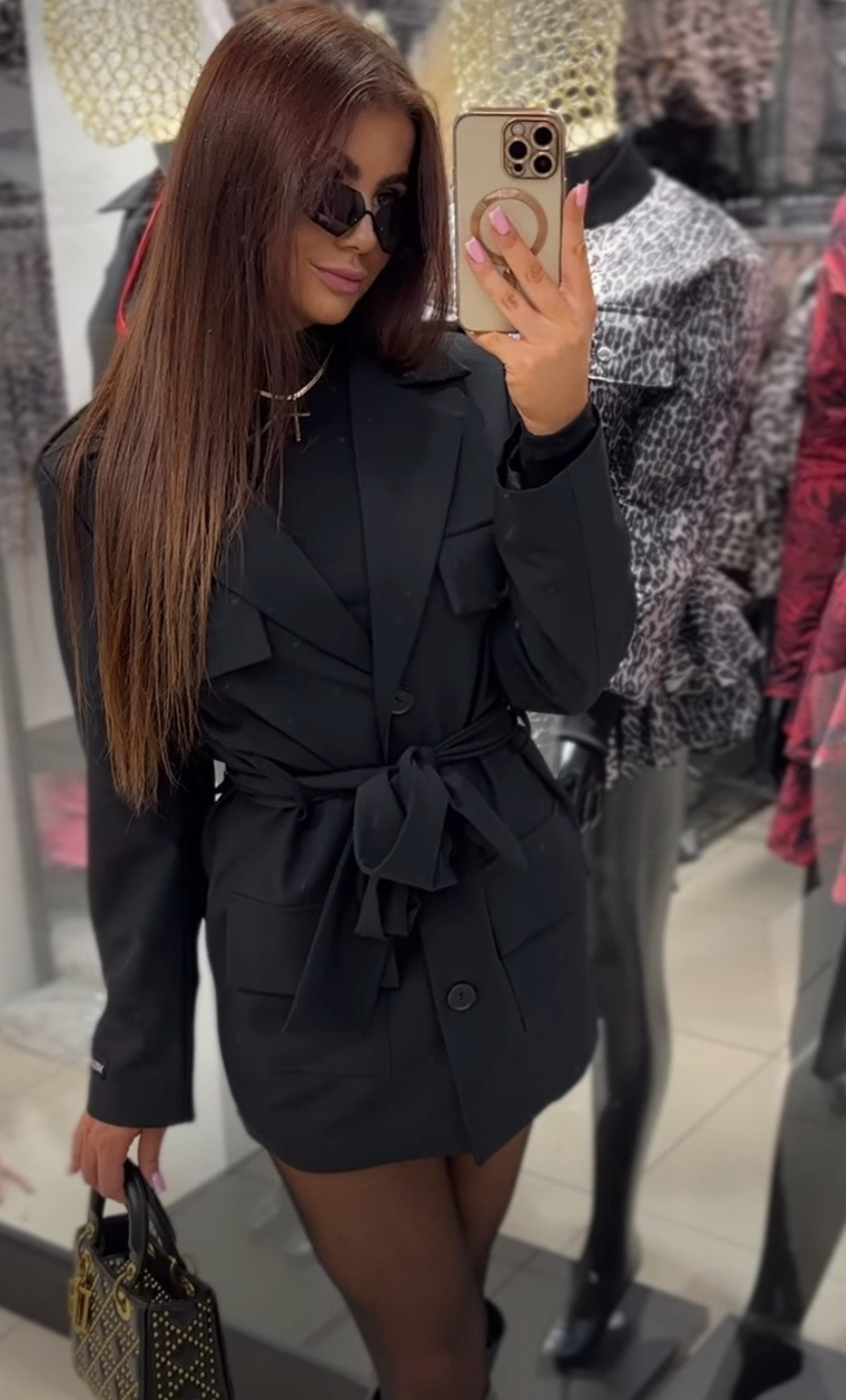 Brunetka w czarnej sukience-płaszczu z paskiem, okularach i z torebką robi selfie w sklepie odzieżowym