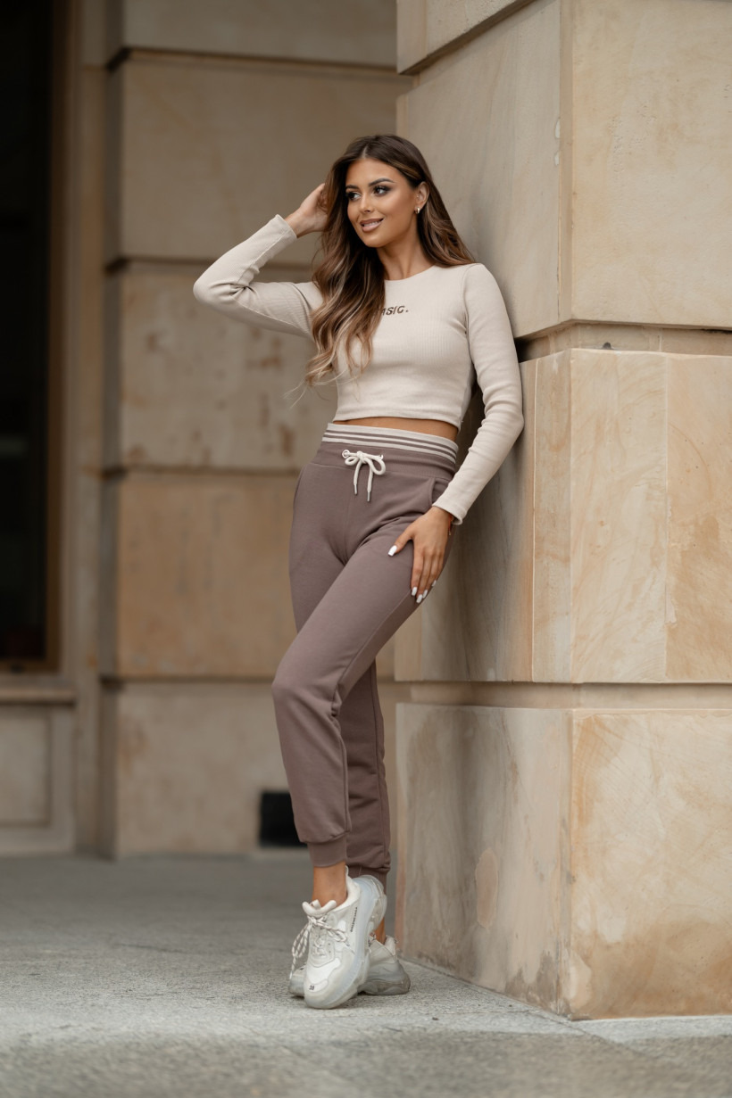 Kobieta opierająca się o kamienny filar w beżowym prążkowanym topie z długim rękawem i brązowych spodniach dresowych ze sznurkiem, do tego białe sportowe buty.