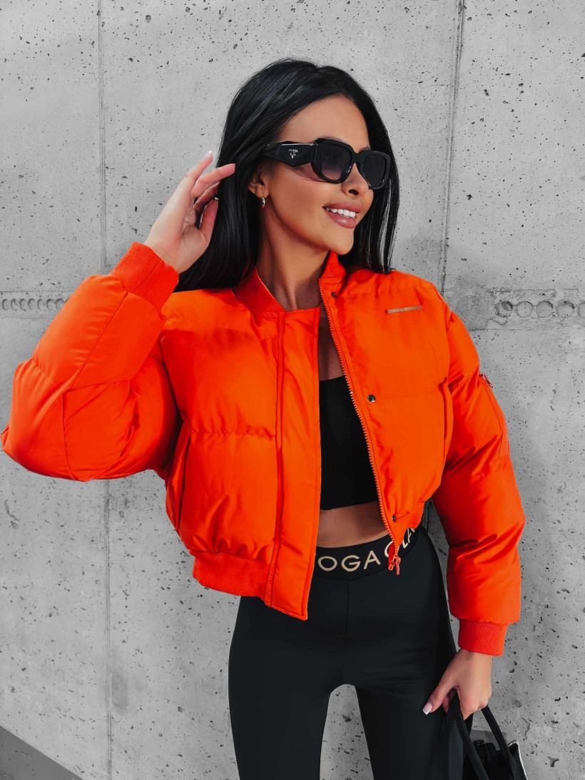 Uśmiechnięta brunetka w czarnych okularach Prada, pomarańczowej kurtce bomberce i czarnych legginsach z napisem, pozuje na tle betonowej ściany.