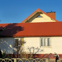 STONEROOF RAFAŁ ZIÓŁKOWSKI - Dom z nowym, czerwonym dachem z blachy trapezowej i rynnami w tym samym kolorze. Widoczne narzędzia dekarskie i fragment elewacji z dwoma oknami.