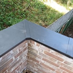 STONEROOF RAFAŁ ZIÓŁKOWSKI - Detal obróbki blacharskiej na murze z cegły, widok z góry na tle zieleni. Precyzyjne wykończenie, szara blacha, mur z czerwonej cegły, zieleń w tle.