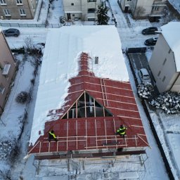 STONEROOF RAFAŁ ZIÓŁKOWSKI - Zimowy widok z góry na częściowo ośnieżony dach z czerwoną membraną i drewnianą konstrukcją. Dwóch dekarzy w żółtych kamizelkach pracuje na rusztowaniu.