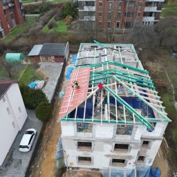 STONEROOF RAFAŁ ZIÓŁKOWSKI - Montaż czerwonej dachówki na nowej więźbie dachowej wielorodzinnego budynku w trakcie budowy, widok z góry na tle zabudowy miejskiej.
