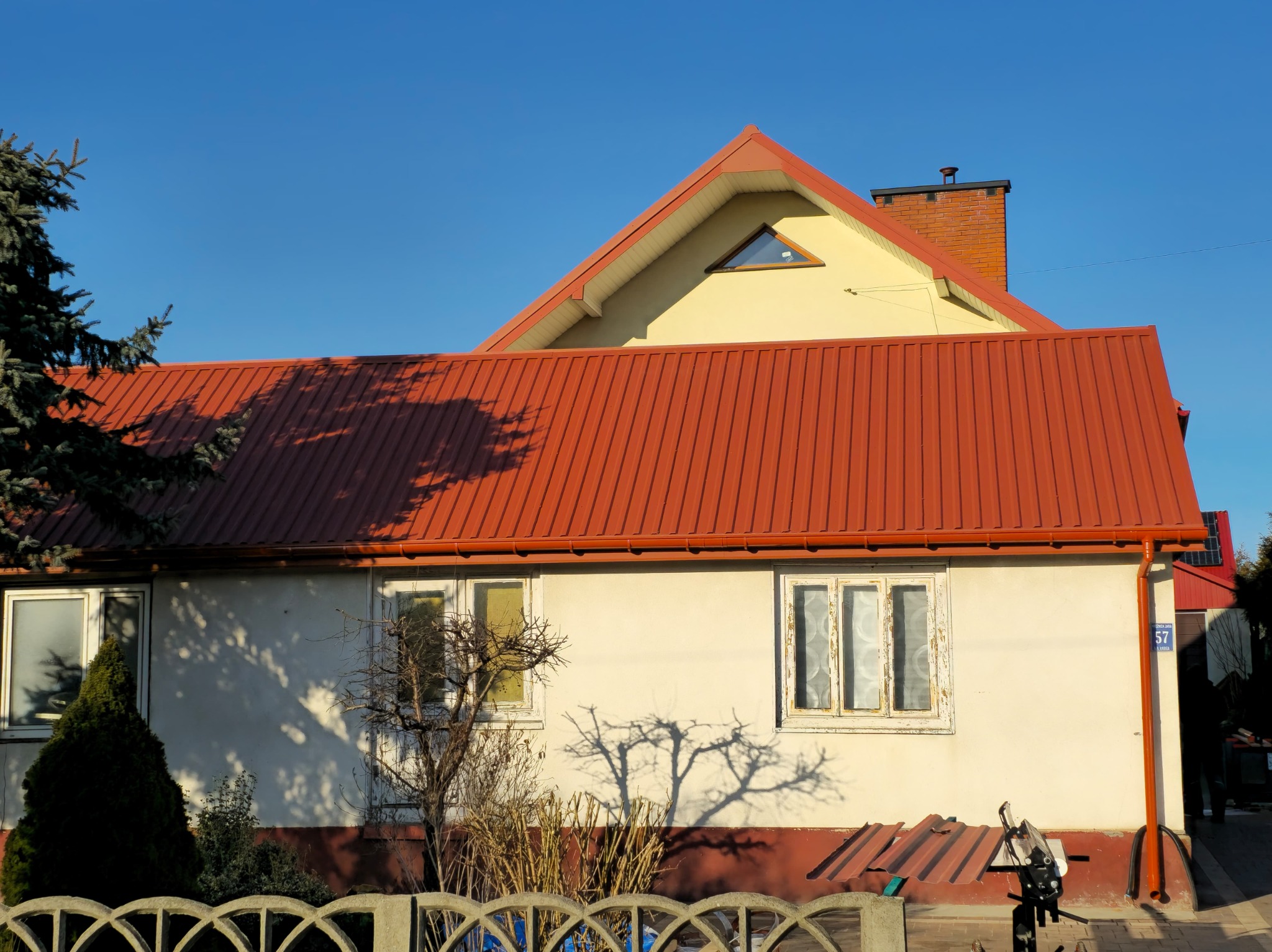 Dom z nowym, czerwonym dachem z blachy trapezowej i rynnami w tym samym kolorze. Widoczne narzędzia dekarskie i fragment elewacji z dwoma oknami.