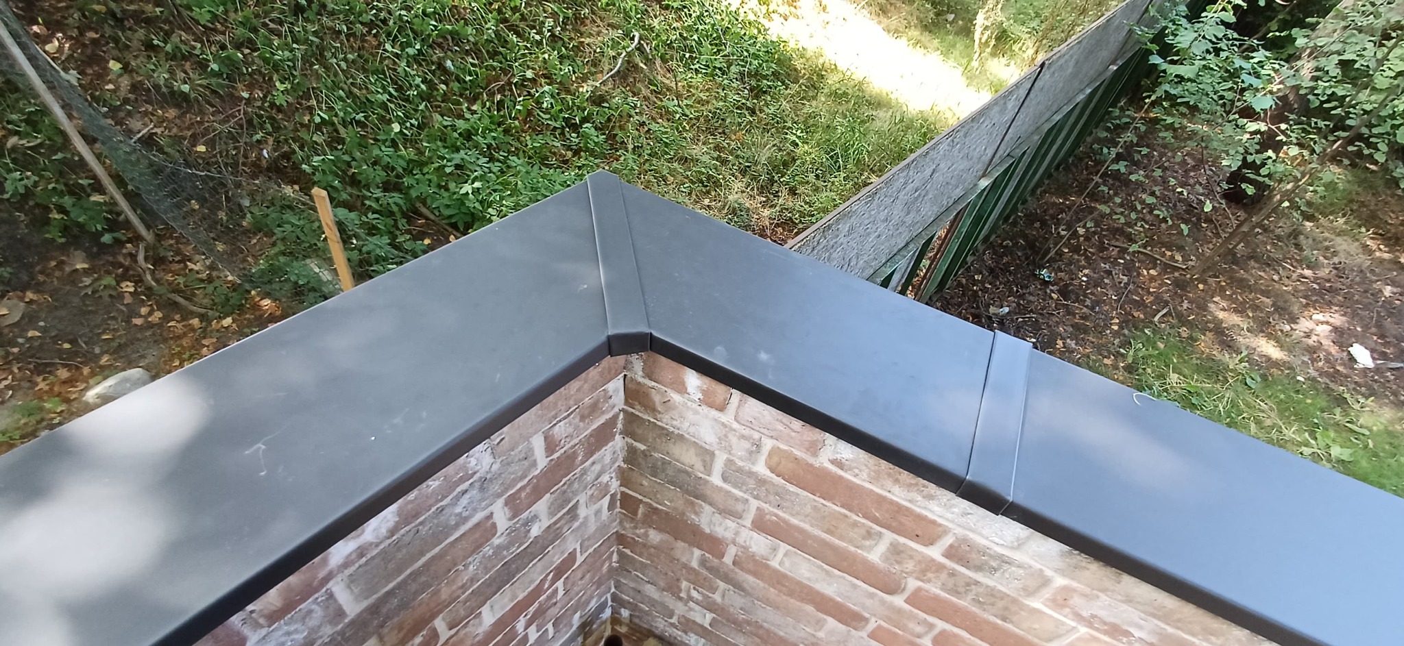 Detal obróbki blacharskiej na murze z cegły, widok z góry na tle zieleni. Precyzyjne wykończenie, szara blacha, mur z czerwonej cegły, zieleń w tle.