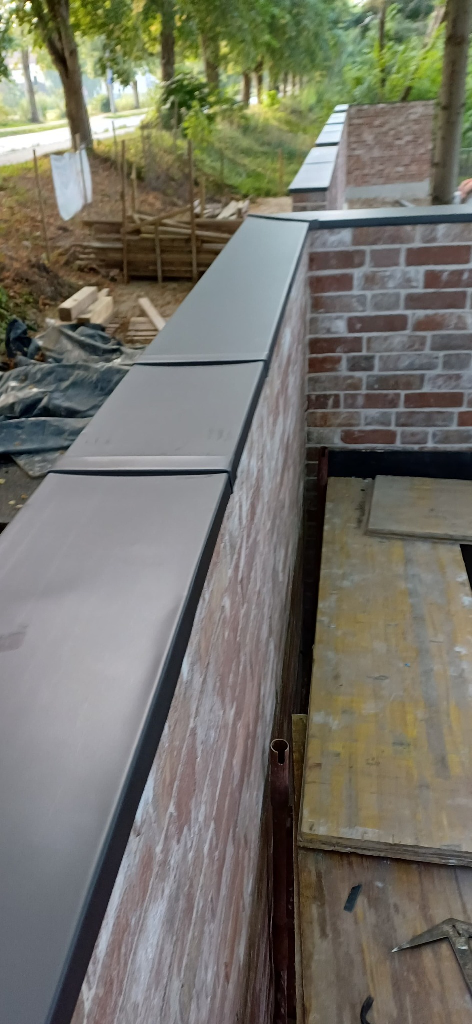 Fragment muru z cegły z zamontowaną, ciemną obróbką blacharską. Widoczne deski i narzędzia w tle. Ujęcie z dołu, lekko zniekształcające perspektywę.