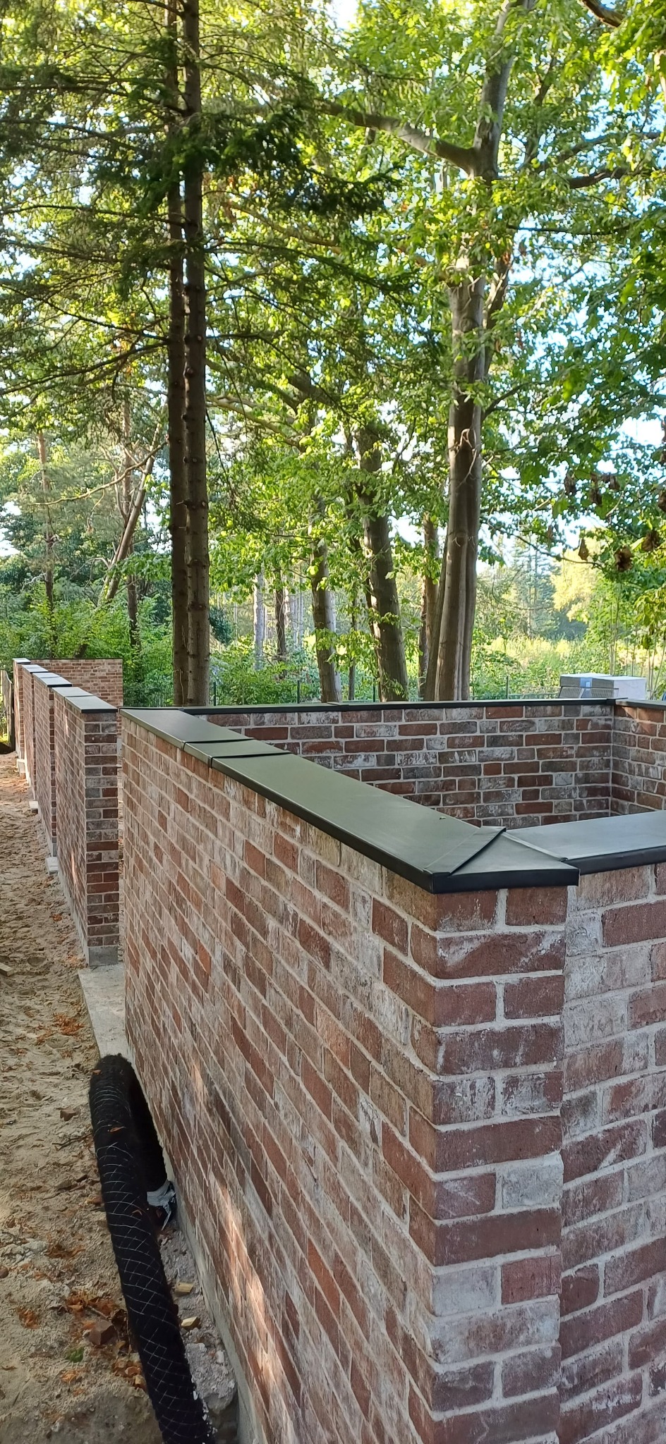 Murowany, ceglany płot z czarnym wykończeniem na tle zielonych drzew. Widok z dołu, ukazujący detale cegieł i otoczenie.