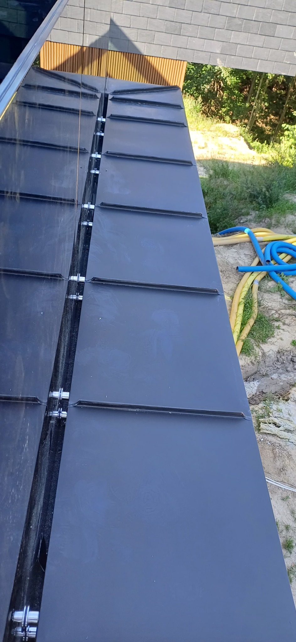 Nowoczesne krycie dachu z blachy na rąbek stojący, widoczne łączenia i mocowania. W tle budynek z drewnianymi elementami elewacji i zieleń.