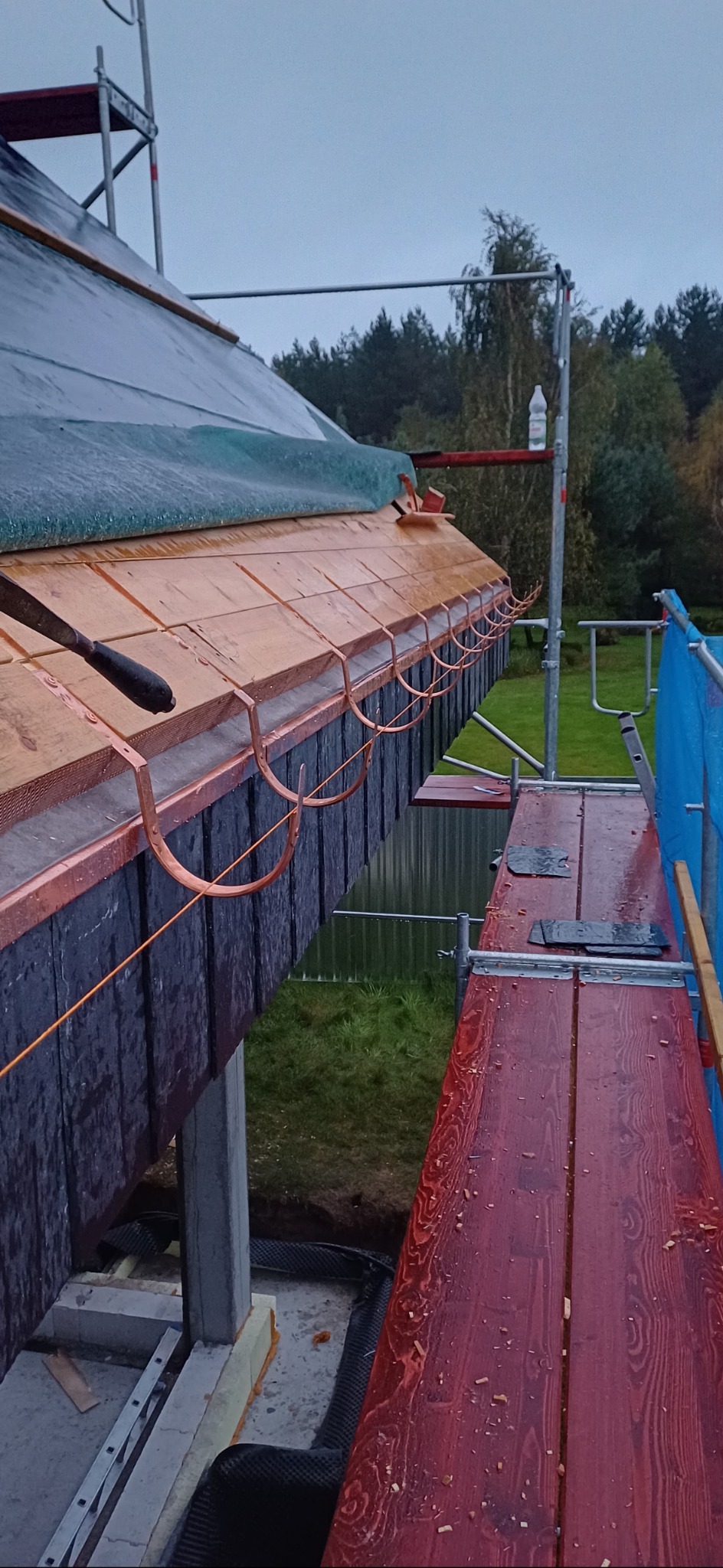 Montaż miedzianych rynien na drewnianej podbitce dachu, widoczne rusztowania i narzędzia. Prace dekarskie w toku, z bliska.