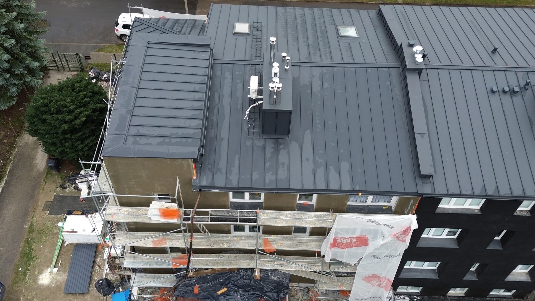 Szary dach z blachy na budynku wielorodzinnym, widok z góry. Na dachu kominy i okna dachowe. Przy ścianie rusztowanie. Prace dekarskie w toku.