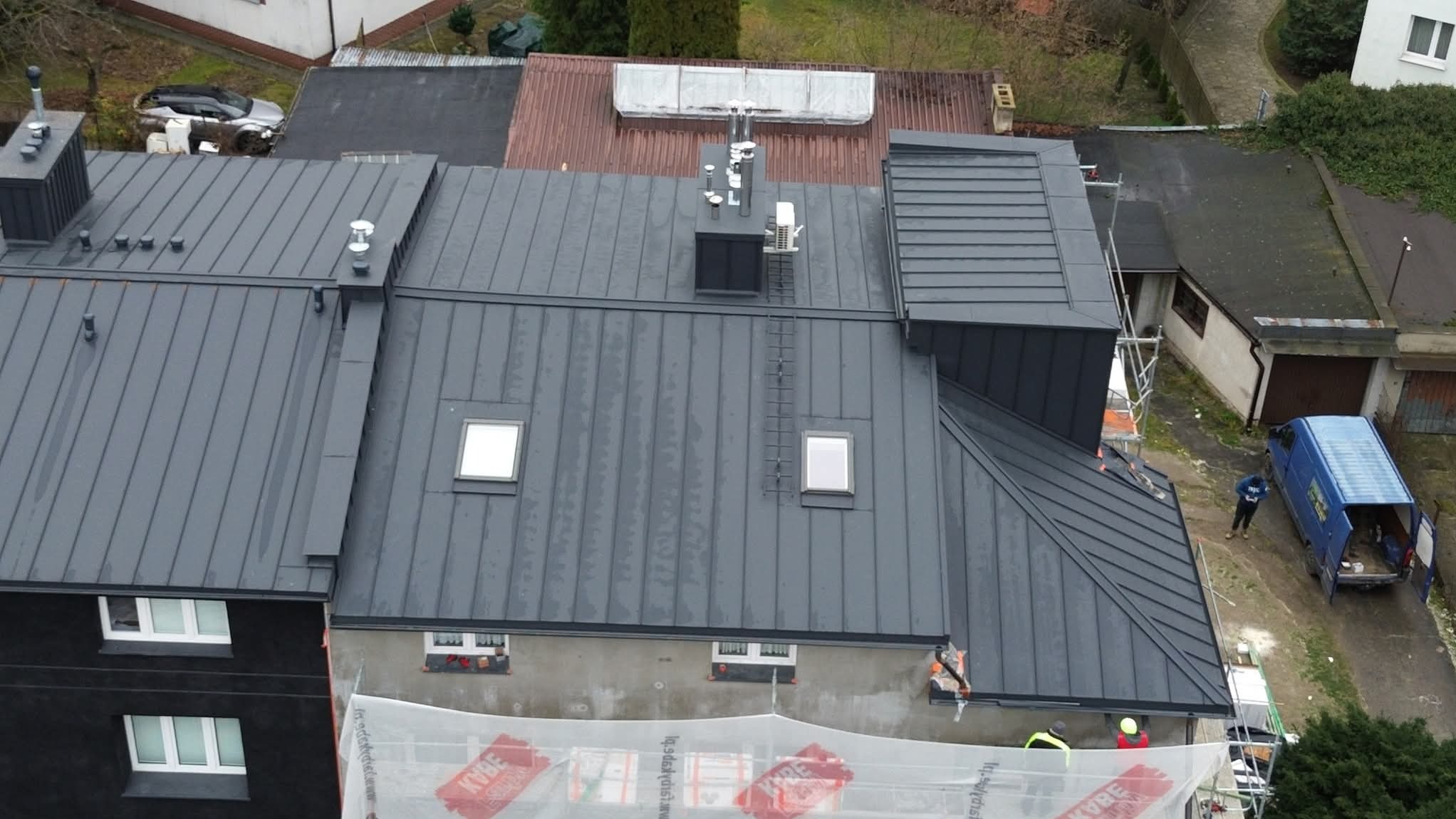 Szary, blaszany dach z oknami dachowymi i kominami, widziany z góry. Na dole rusztowania i siatka ochronna. W tle zabudowa jednorodzinna i zaparkowany niebieski bus.