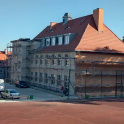 Budynek w trakcie prac elewacyjnych, widoczne rusztowania, częściowo ocieplona i pomalowana fasada, zaparkowane samochody dostawcze i osobowe, boisko sportowe na pierwszym planie.