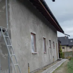 Elewacja budynku w trakcie tynkowania maszynowego, widoczne rusztowanie i zabezpieczone okna folią, zacierana faktura tynku.