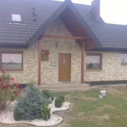 Dom jednorodzinny z elewacją wykończoną kamieniem dekoracyjnym, drewnianym zadaszeniem nad wejściem i zadbanym ogrodem z różami i iglakami.