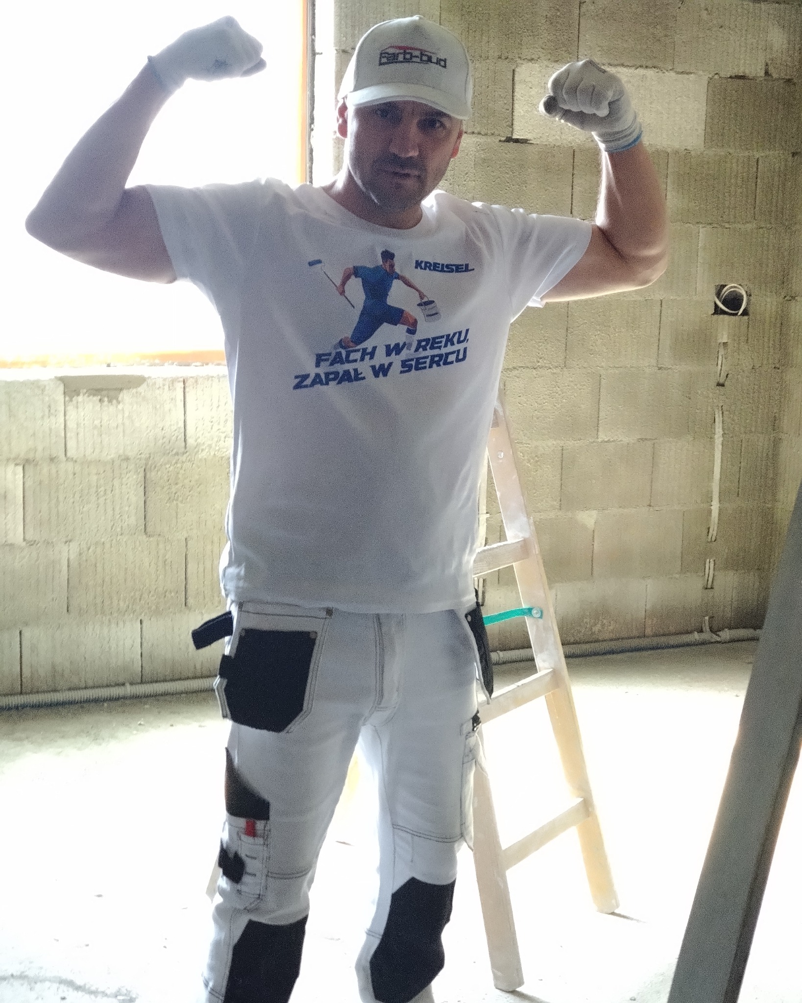 Mężczyzna w roboczym stroju, koszulce z nadrukiem i czapce z daszkiem, prezentuje siłę na tle surowych ścian z betonu komórkowego i drewnianej drabiny.