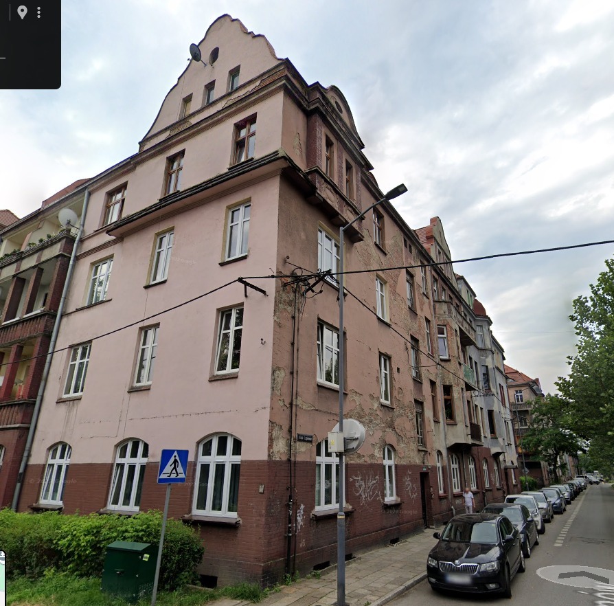 Przedwojenna kamienica z widocznymi śladami uszkodzeń elewacji, zlokalizowana przy ulicy z zaparkowanymi samochodami i zielenią. Widoczne okna i detale architektoniczne.