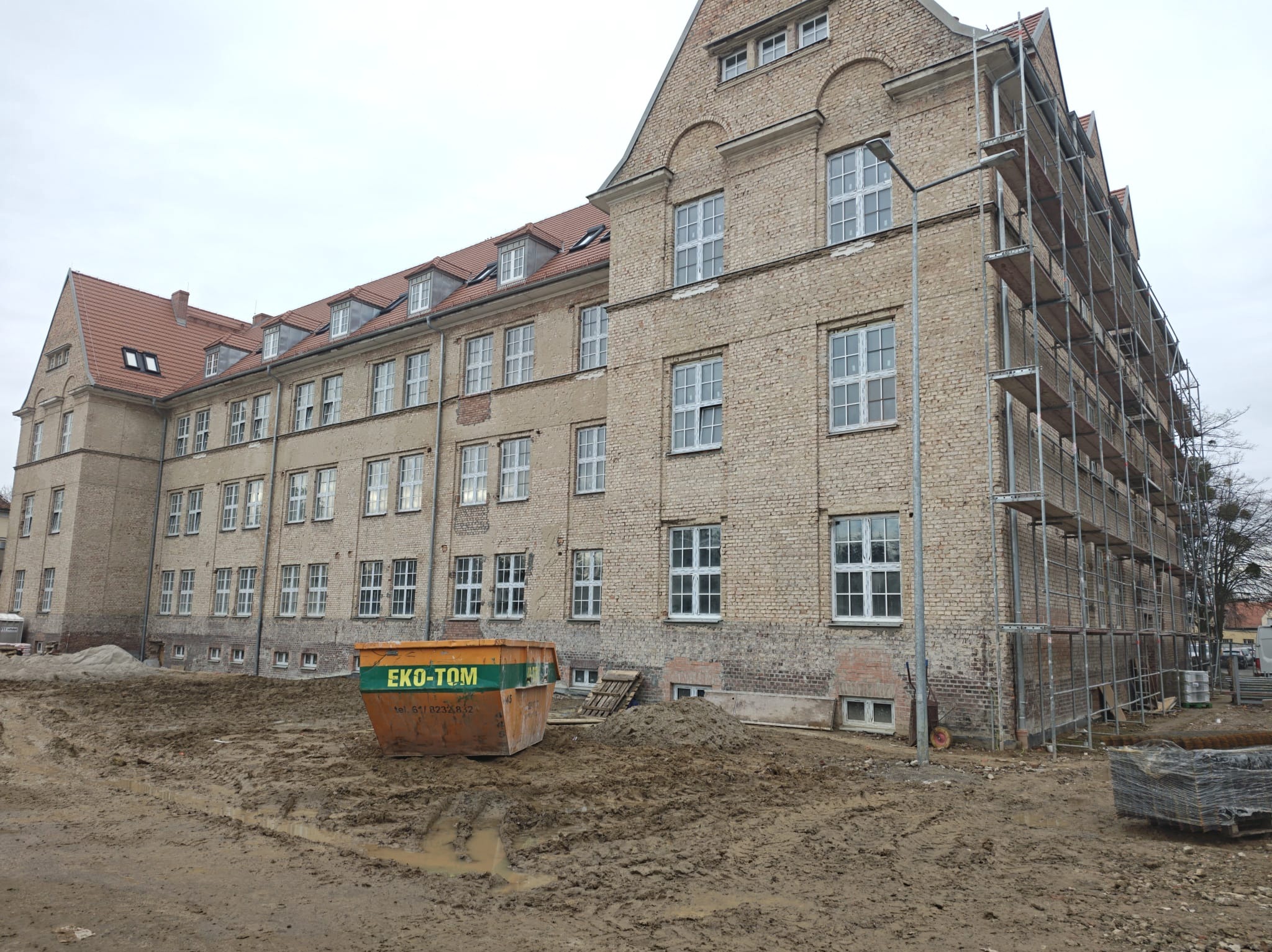 Budynek z cegły w trakcie renowacji, otoczony rusztowaniami i kontenerem na gruz. Teren wokół rozkopany, widoczne ślady błota i piasku. Pochmurne niebo w tle.