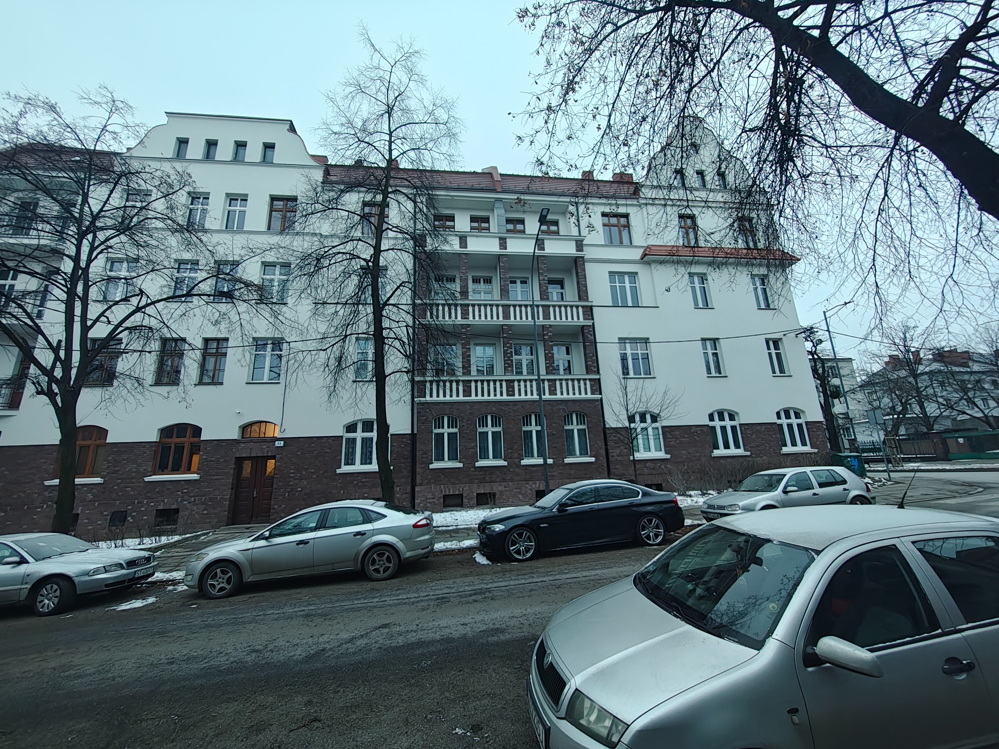 Odnowiona kamienica z białymi ścianami i balkonami, widoczna z poziomu ulicy z zaparkowanymi samochodami. Uwagę zwraca odświeżona elewacja budynku.