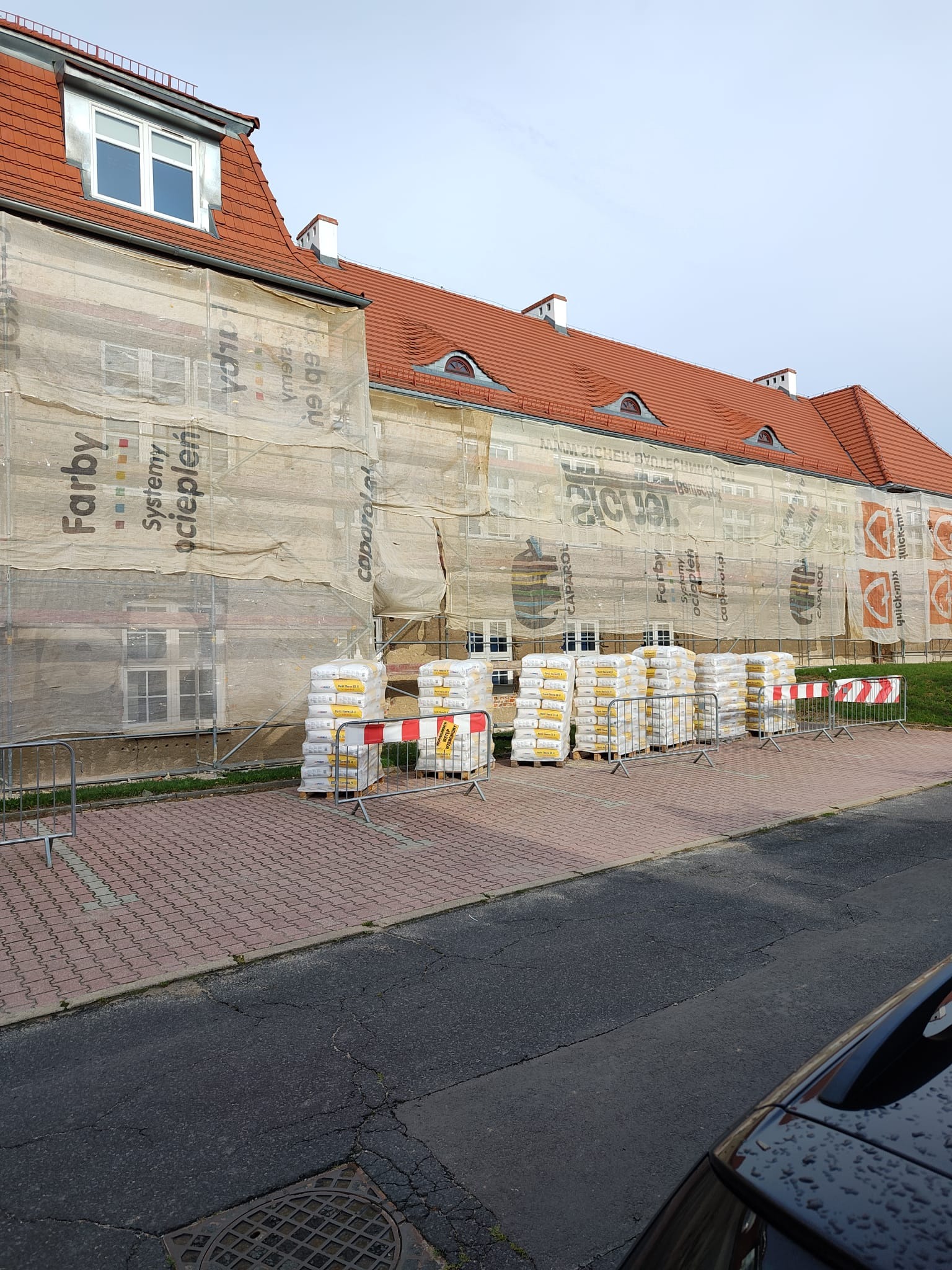 Budynek w trakcie prac elewacyjnych, osłonięty siatką z nadrukiem reklamowym, palety z materiałami budowlanymi zabezpieczone barierkami.
