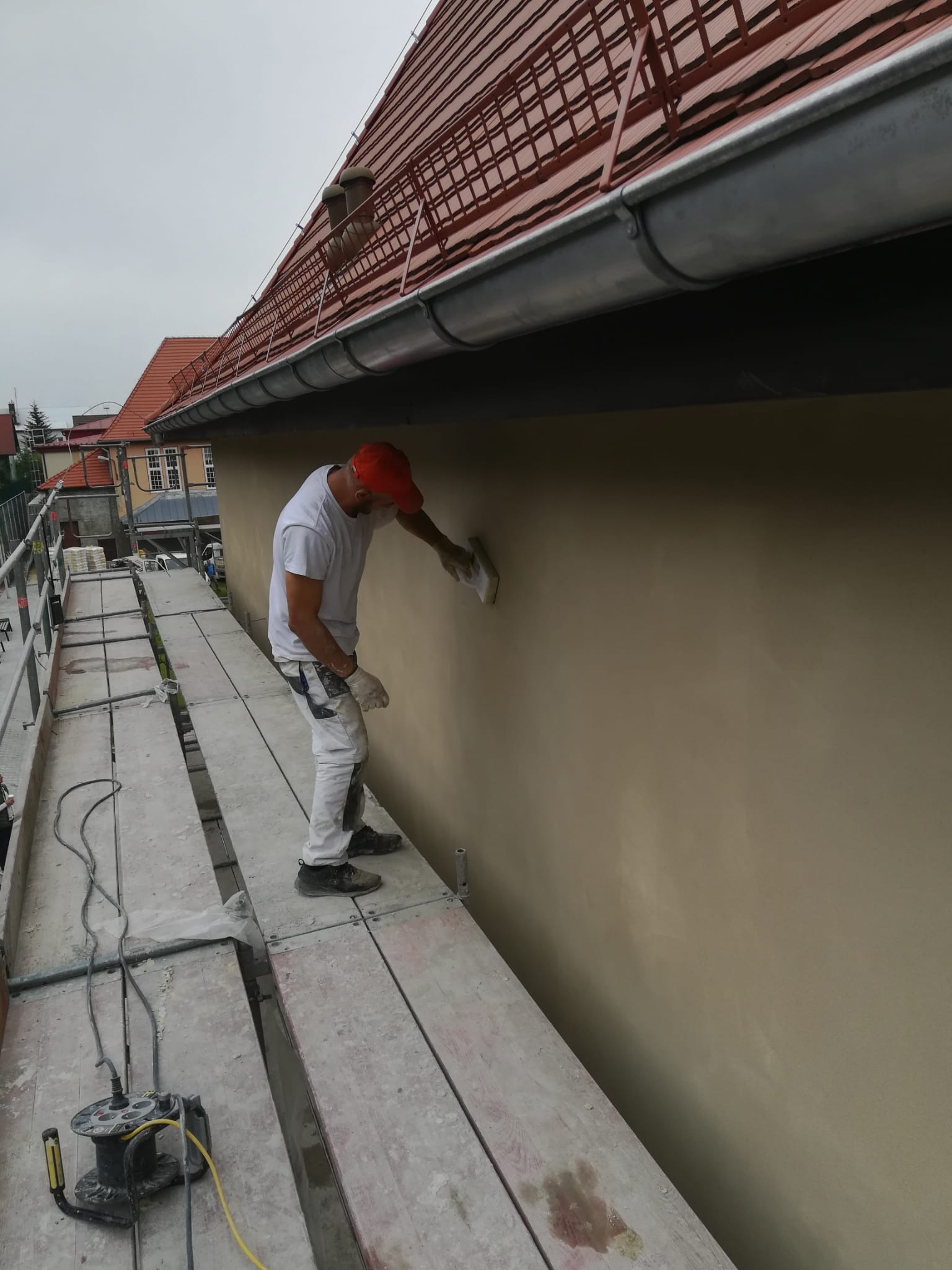 Pracownik w czerwonej czapce wygładza szpachelką świeżo nałożony tynk na elewacji domu, stojąc na rusztowaniu; widoczny zwinięty przedłużacz elektryczny.