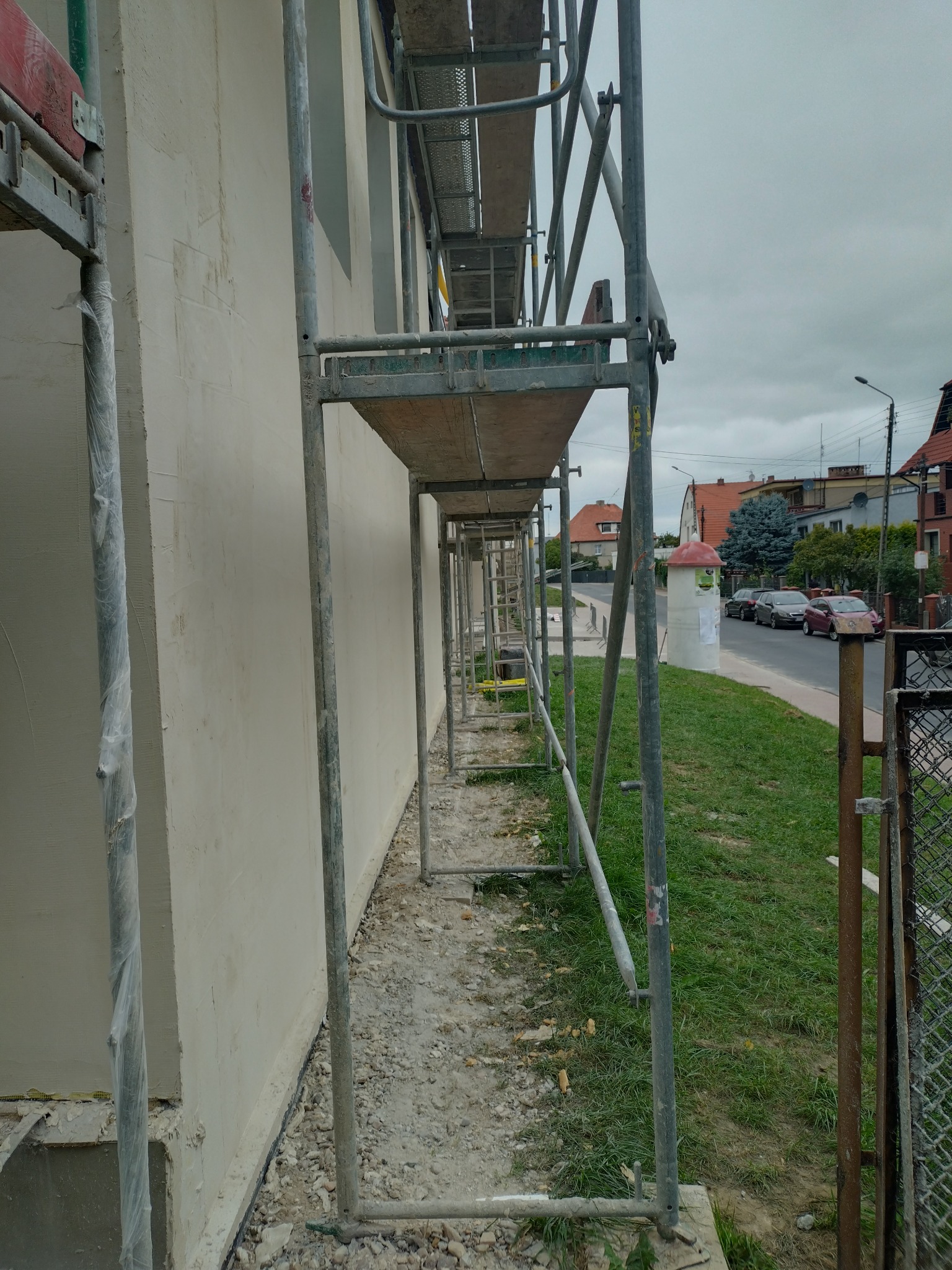 Świeżo otynkowana elewacja budynku w trakcie schnięcia, widoczna spod rusztowania, z fragmentem ulicy w tle.