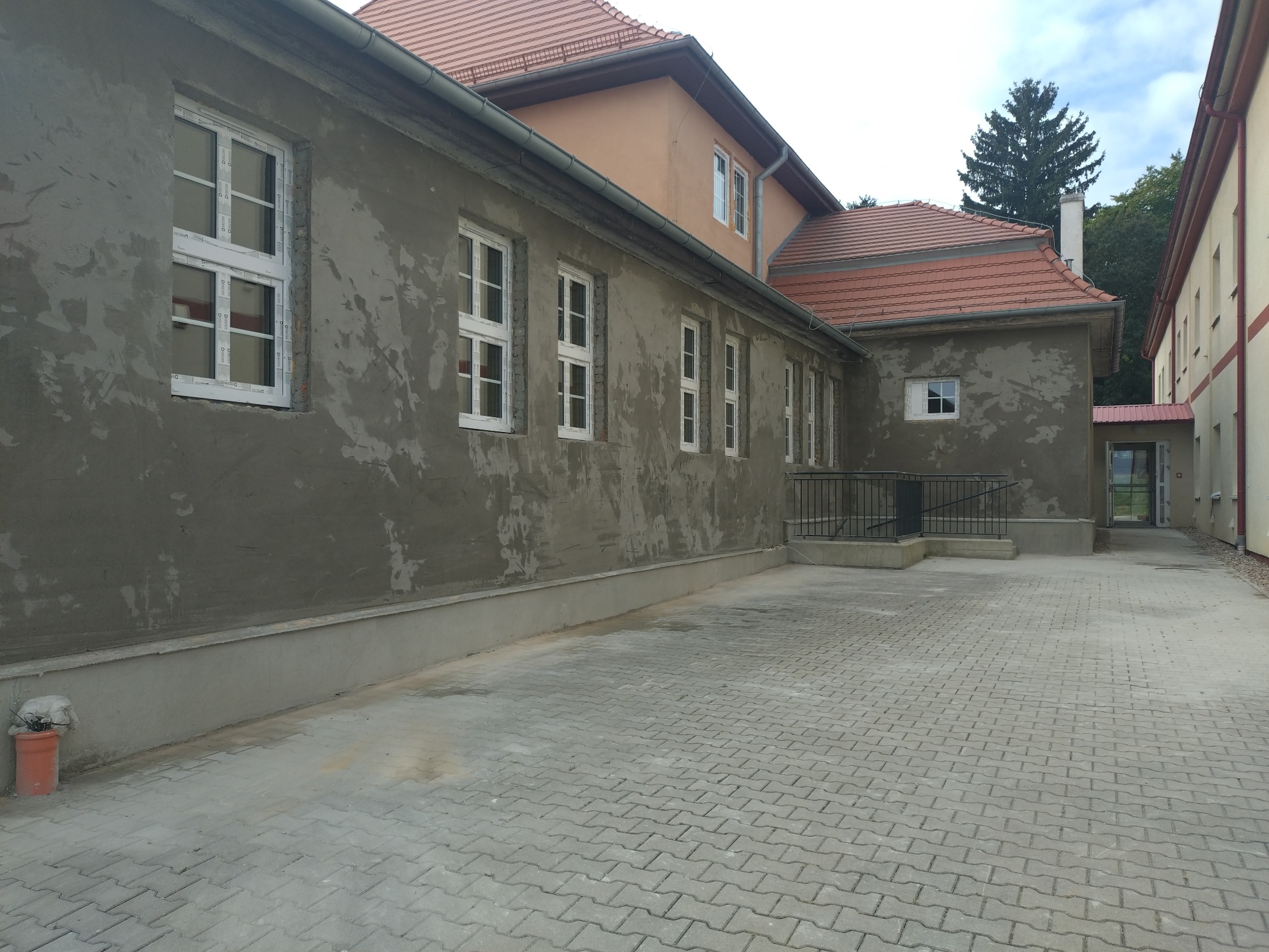 Elewacja budynku w trakcie prac tynkarskich, widoczne surowe tynki i nowe okna PCV, kostka brukowa na dziedzińcu.