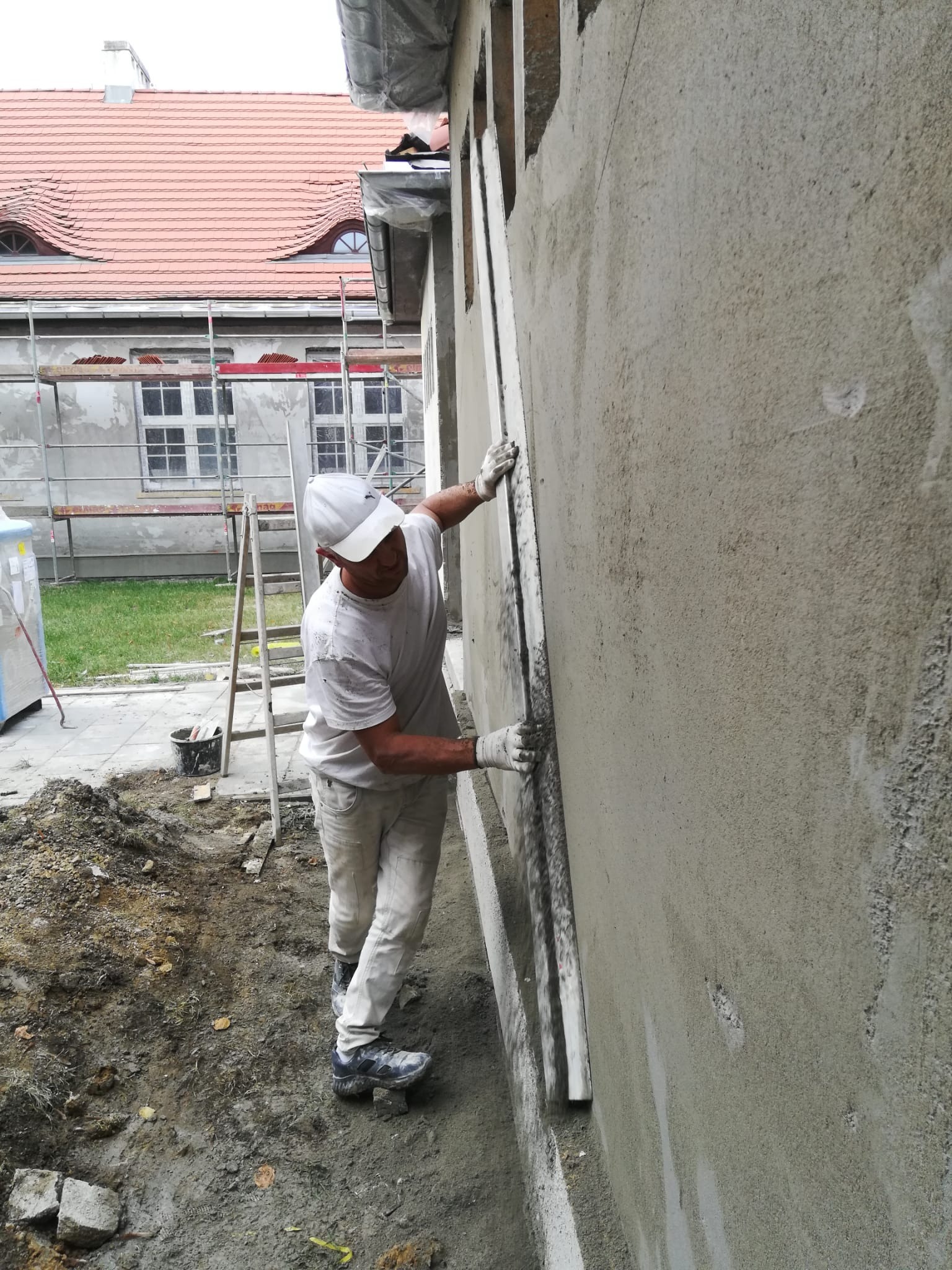 Pracownik w białym ubraniu wyrównuje świeżo nałożony tynk na elewacji budynku za pomocą długiej łaty, widoczne rusztowania i fragment dachu z lukarnami w tle.