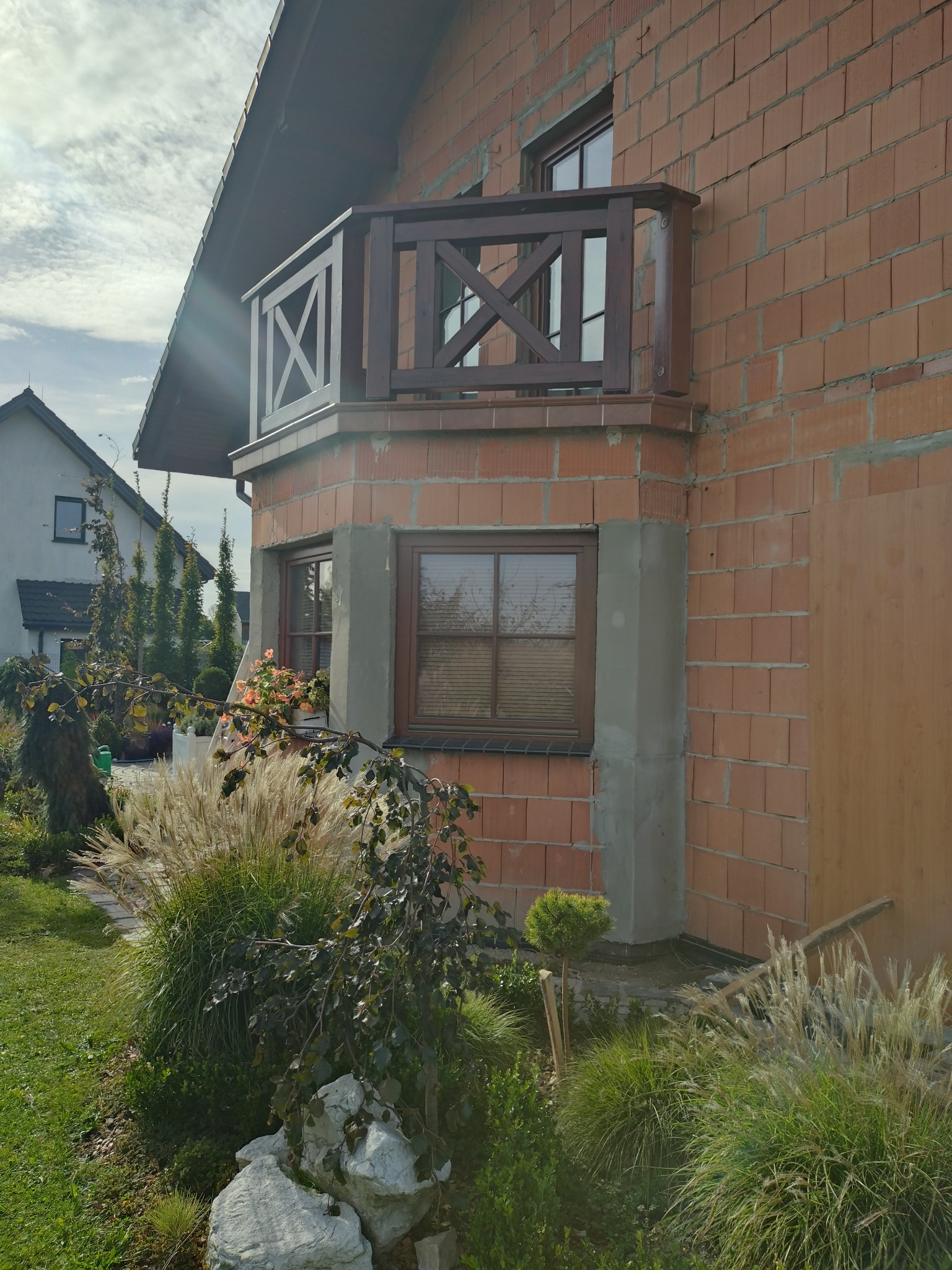 Surowa elewacja budynku z czerwonej cegły z drewnianym balkonem, oknami i częściowo otynkowanymi narożnikami, otoczona zielenią i elementami ogrodu.
