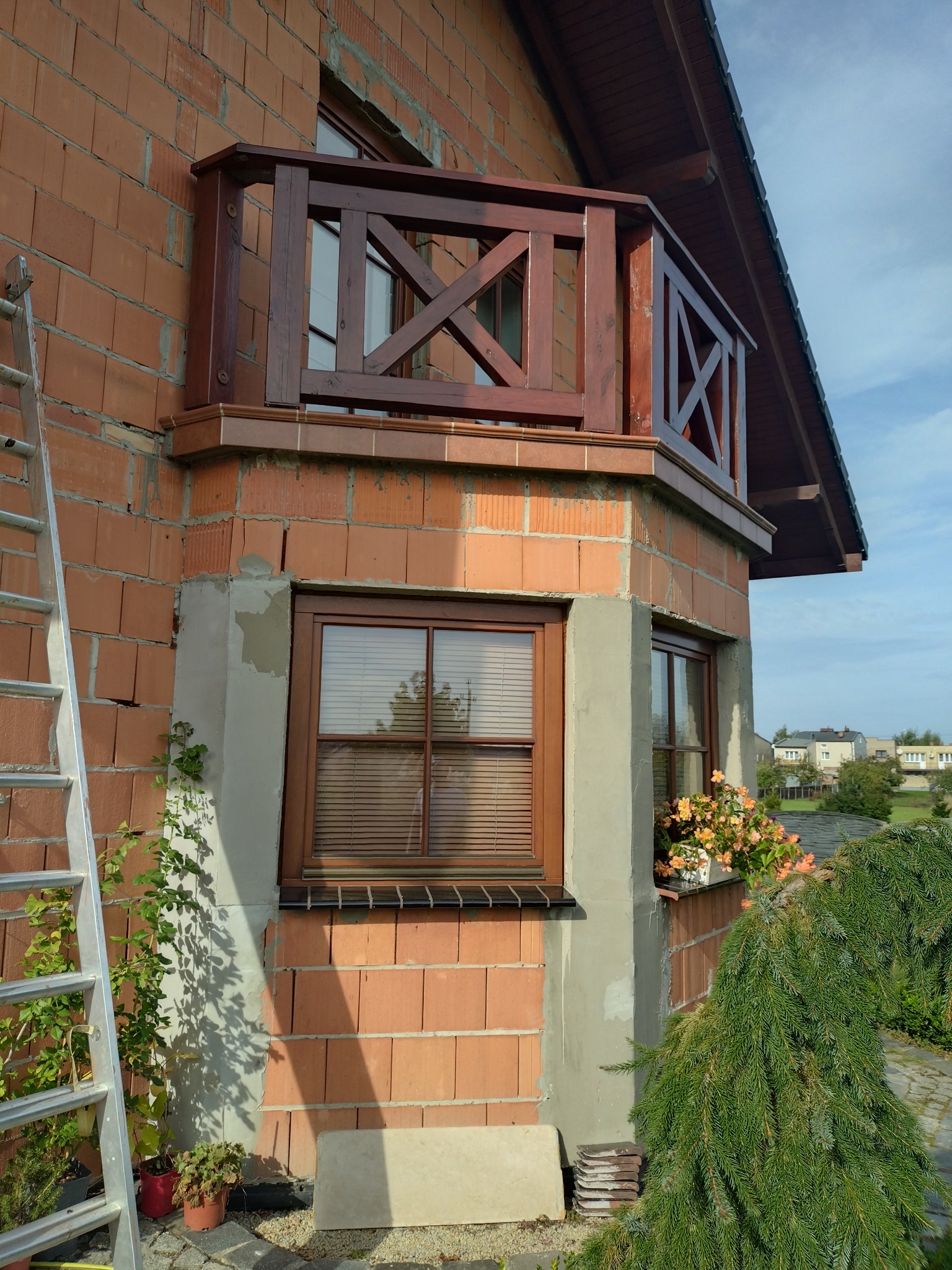 Fragment elewacji domu z balkonem, widoczny fragment ściany z czerwonej cegły z oknem, częściowo pokryty szarym tynkiem, aluminiowa drabina oparta o ścianę. Na balkonie drewniana balustrada...