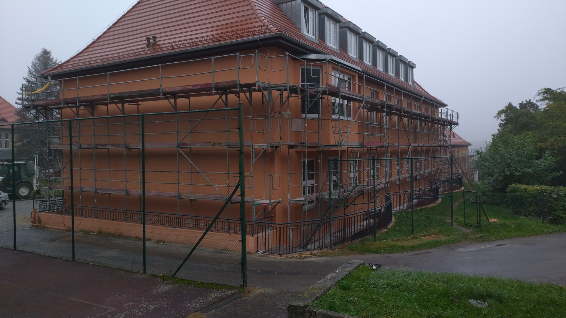 Budynek wielopiętrowy w trakcie prac elewacyjnych, otoczony rusztowaniem, widoczny traktor w tle, pochmurny dzień.