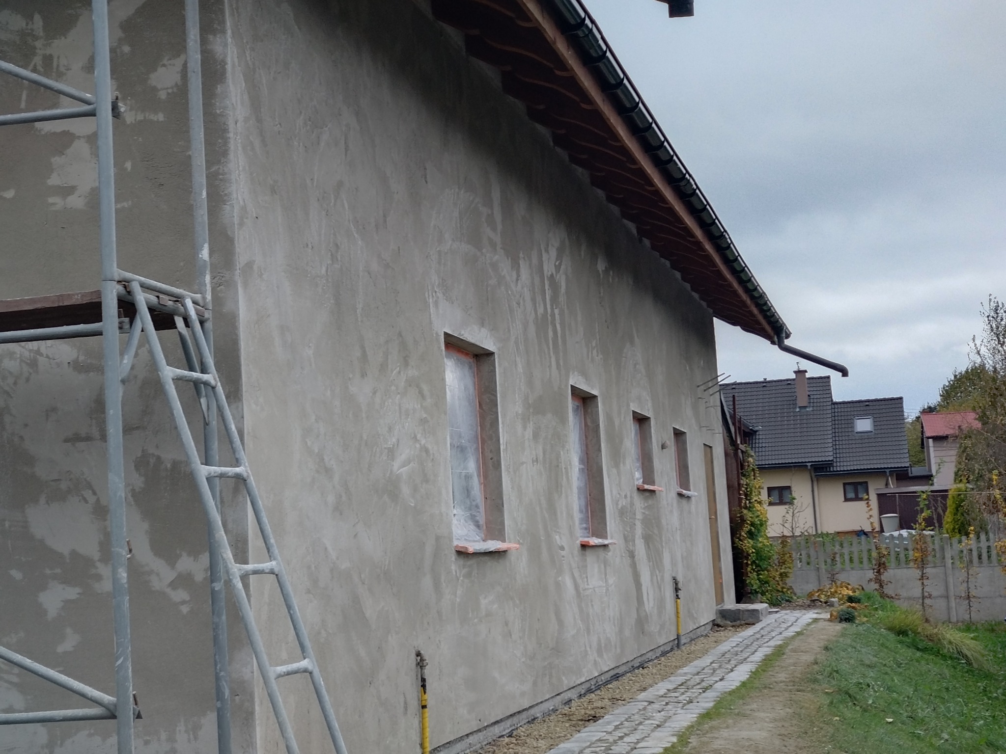 Elewacja budynku w trakcie tynkowania maszynowego, widoczne rusztowanie i zabezpieczone okna folią, zacierana faktura tynku.