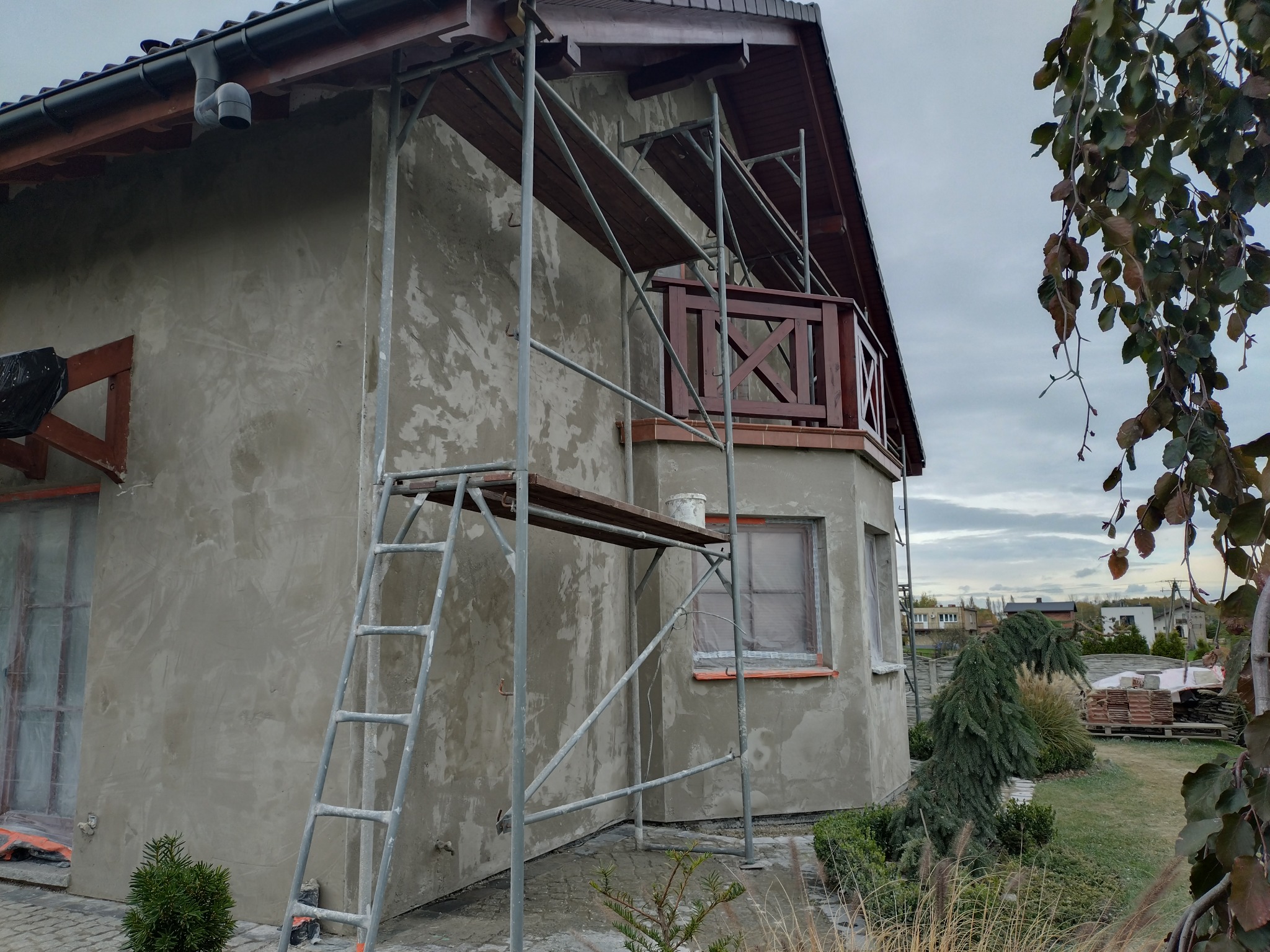 Dom jednorodzinny w trakcie prac elewacyjnych: ściany pokryte surowym tynkiem, metalowe rusztowanie, okna zabezpieczone folią, widoczny fragment ogrodu z drzewami i krzewami.