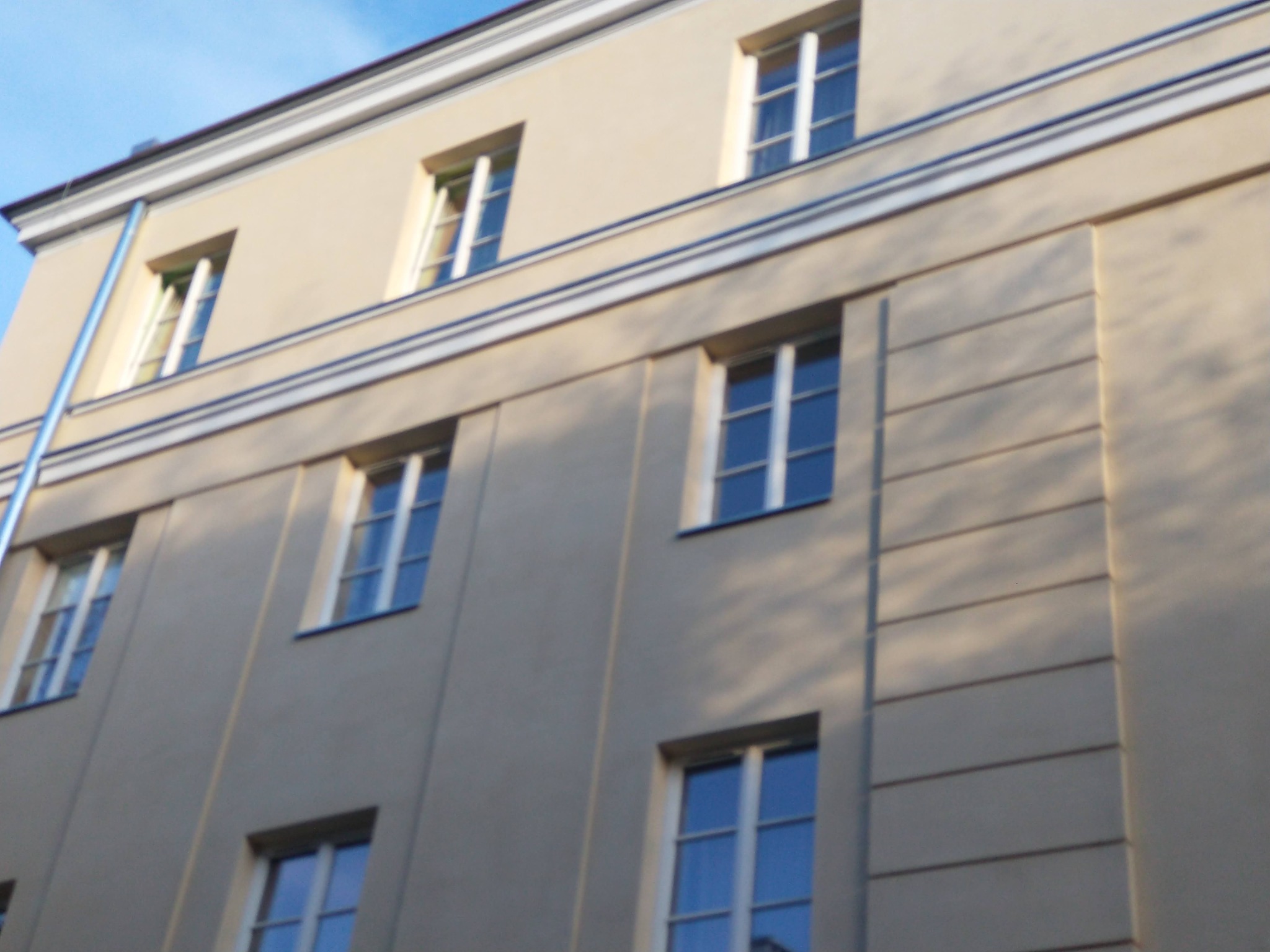 Ocieplona elewacja budynku wielorodzinnego z kremową fasadą, widoczne okna z białymi ramami, dekoracyjne listwy i rynna, ujęcie od dołu w słoneczny dzień.