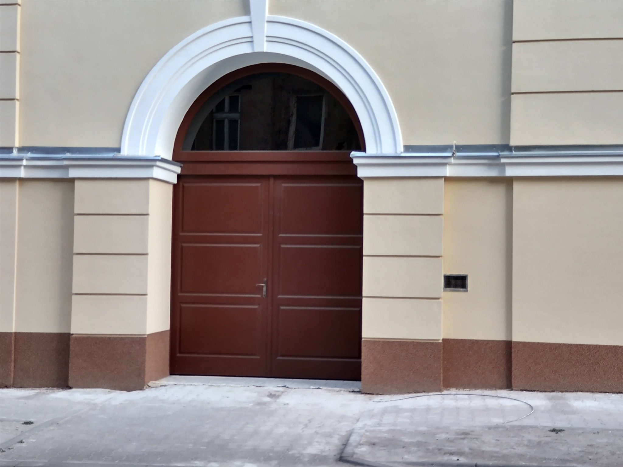 Elewacja budynku z brązowymi dwuskrzydłowymi drzwiami i półokrągłym, białym nadprożem. Dolna część ściany wykończona w kolorze brązowym, reszta w odcieniach beżu.