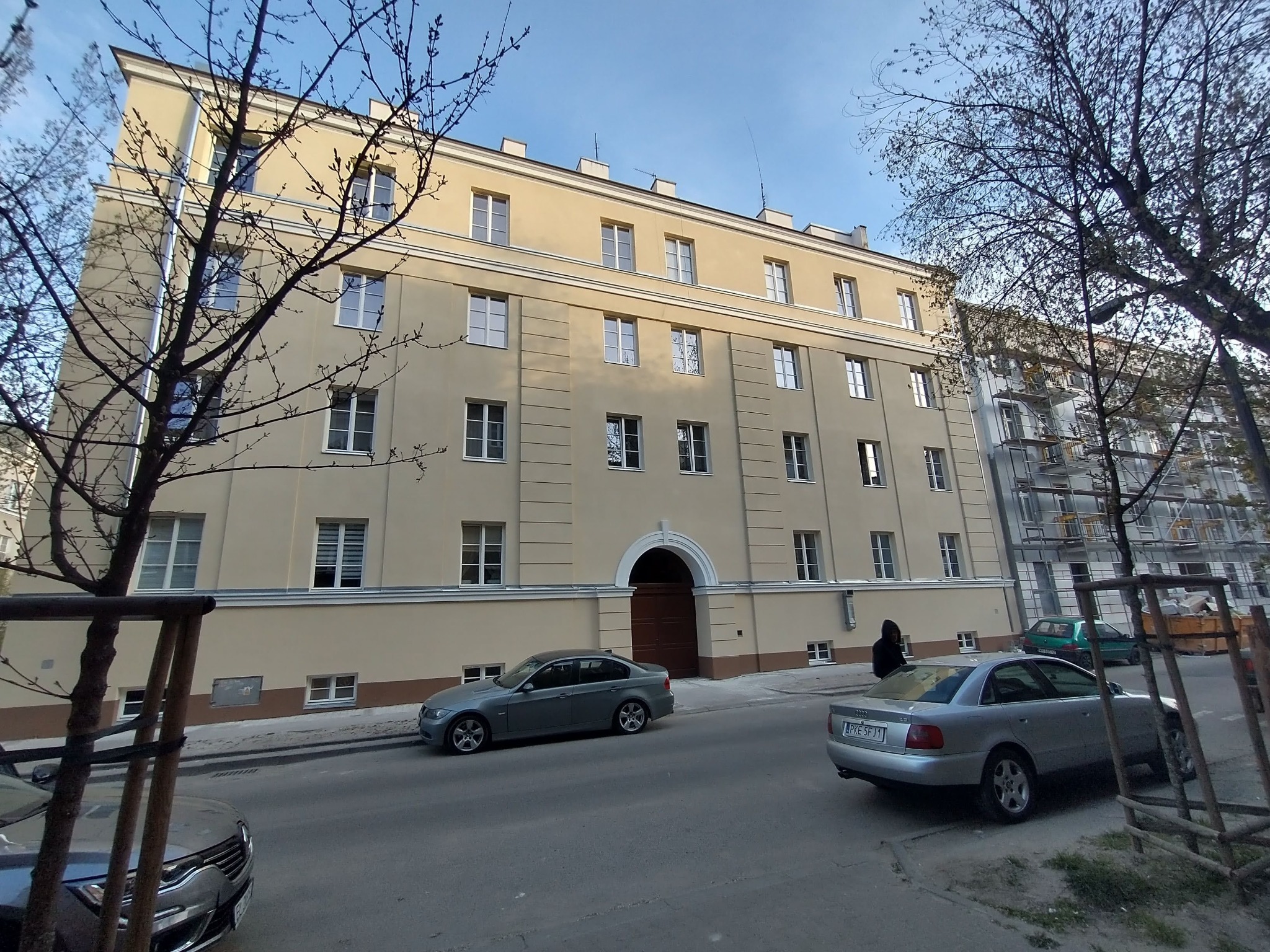 Elewacja odnowionego czteropiętrowego budynku z beżową fasadą i białymi oknami, widoczna brama łukowa, zaparkowane samochody na ulicy, drzewa bez liści na pierwszym planie, rusztowania na sąsiednim...