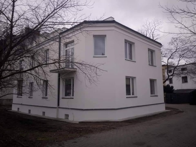 Dwupiętrowy budynek mieszkalny z balkonem po remoncie elewacji, widoczny z perspektywy narożnej, drzewa bez liści w tle.