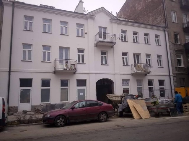 Odnowiona fasada trzypiętrowej kamienicy w jasnych kolorach, z balkonami o metalowych balustradach, widoczny samochód osobowy i sprzęt budowlany na pierwszym planie, sugerujący trwające prace...