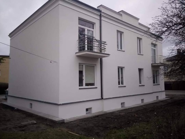 Dwupiętrowy budynek mieszkalny z jasną elewacją i balkonami z metalowymi balustradami, widok z zewnątrz, z widocznym fragmentem ogrodu i drzewa bez liści.