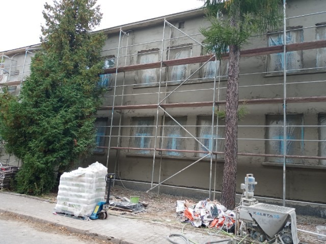 Budynek wielorodzinny w trakcie prac elewacyjnych: ściany pokryte szarym tynkiem, okna zabezpieczone folią, rusztowanie, paleta z workami materiałów budowlanych i agregat tynkarski Kaleeta 5S...