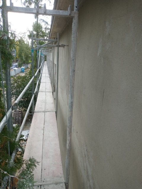 Elewacja budynku w trakcie prac wykończeniowych, widoczne rusztowania i świeżo nałożony tynk o szarej barwie.