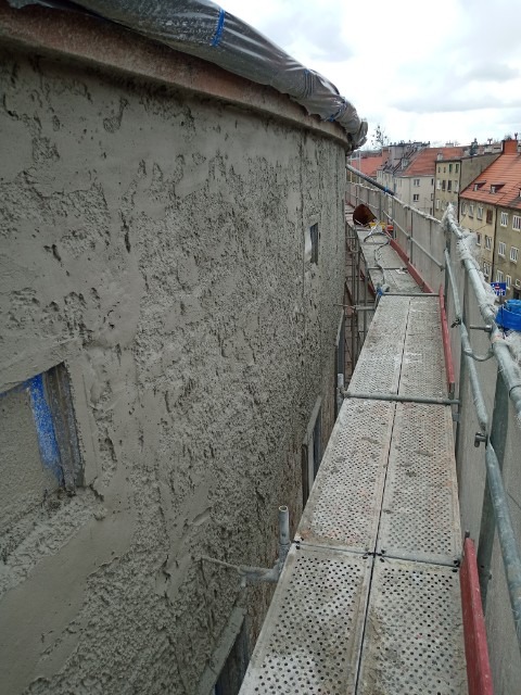 Ściana budynku pokryta świeżym, surowym tynkiem, widoczna z poziomu rusztowania, z fragmentami okien w tle i zadaszeniem zabezpieczonym folią na górze.