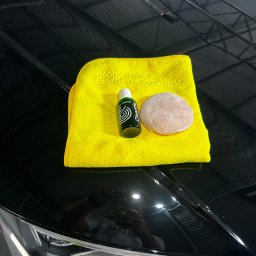 BM GARAGE - Żółta ściereczka mikrofibra, aplikator i butelka środka do pielęgnacji lakieru na błyszczącej, czarnej masce samochodu.