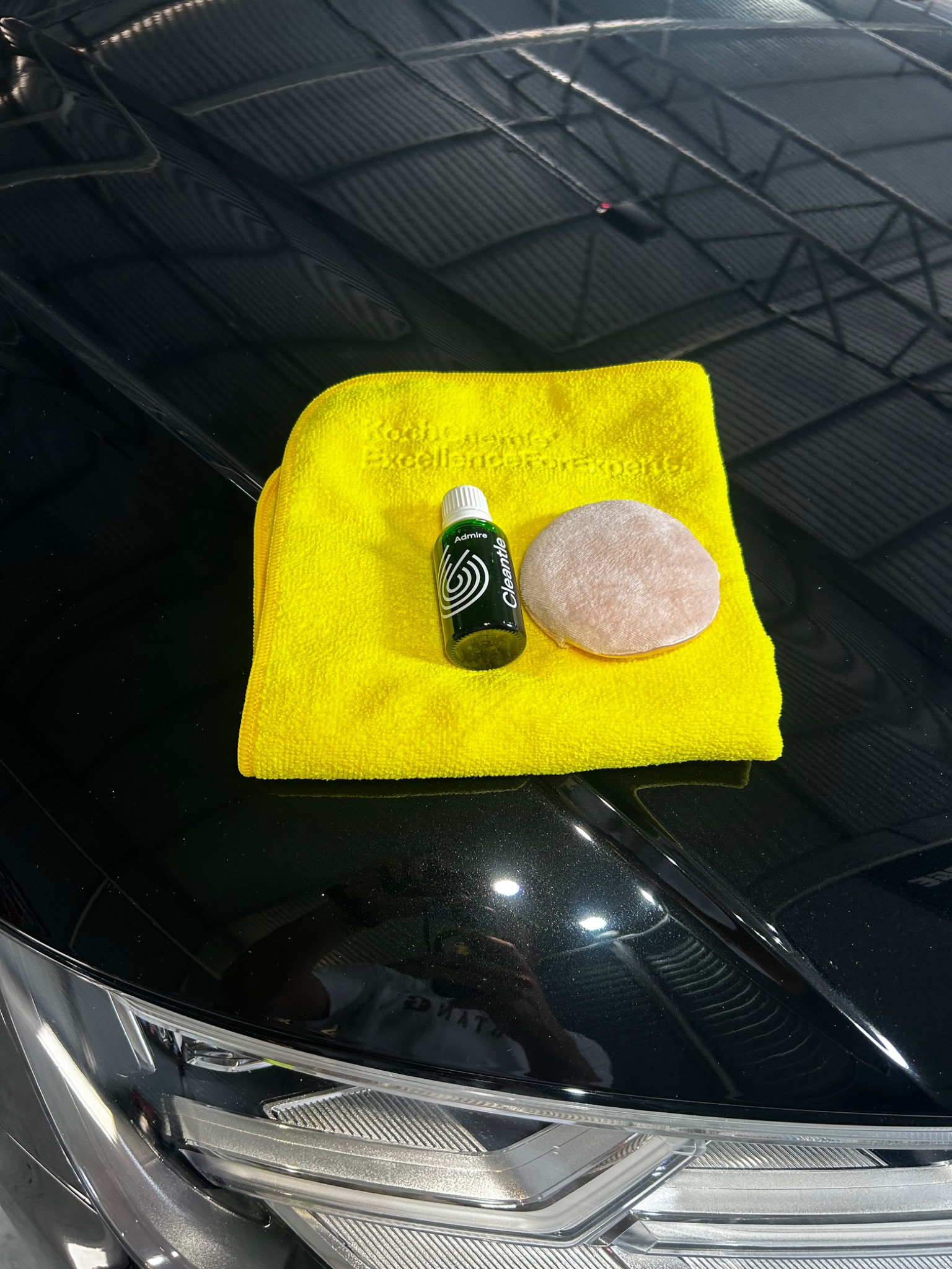 Żółta ściereczka mikrofibra, aplikator i butelka środka do pielęgnacji lakieru na błyszczącej, czarnej masce samochodu.