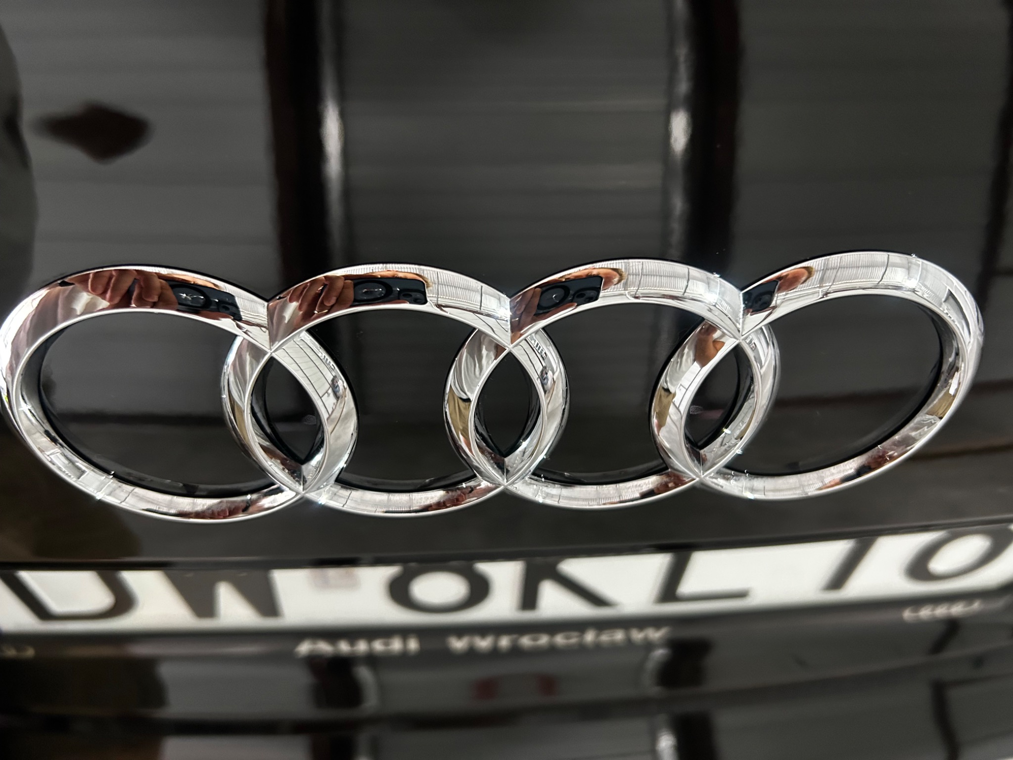 Chromowane logo Audi na czarnym lakierze samochodu, z widocznym fragmentem tablicy rejestracyjnej i odbiciem otoczenia w lakierze.